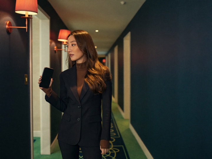 A woman in a tailored blazer in a hotel corridor at night, holding a phone at chest height with the screen dark