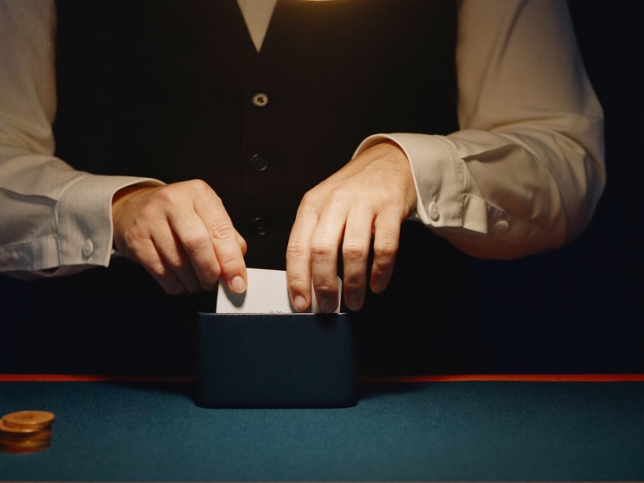 A dealer's hands in a waistcoat and white cuffs drawing a plain card from a card dispenser on dark felt