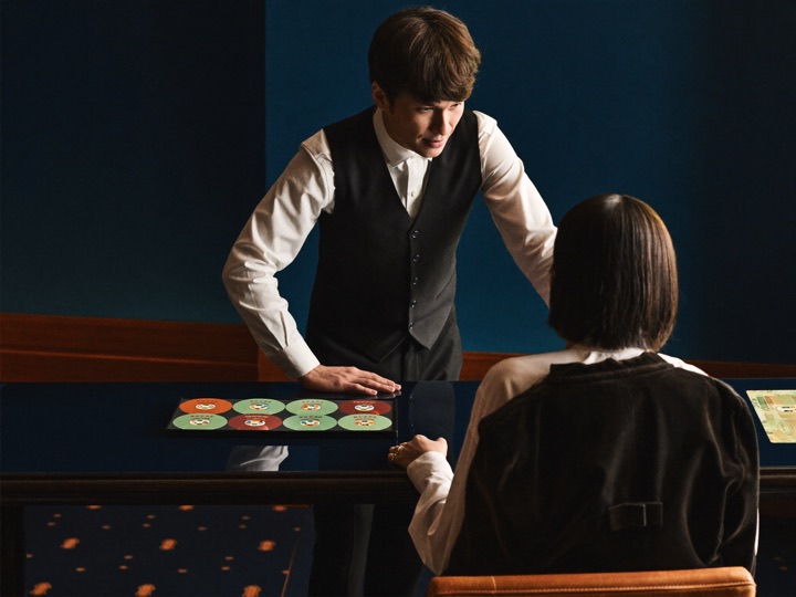 A host in a waistcoat leaning on a gaming counter, talking to a seated guest seen from behind over a row of copper and mint chips