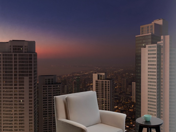 A Kuala Lumpur skyline at dusk seen from a high window, an empty armchair in front