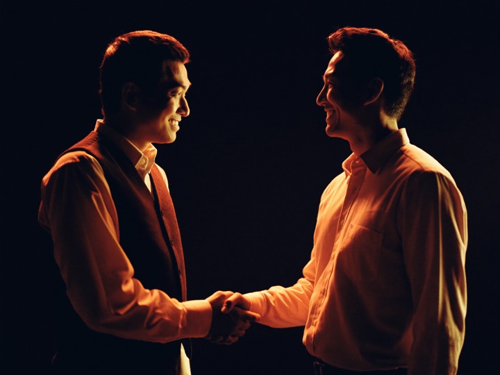 A host in a waistcoat shaking hands with a newly arrived guest against a dark background