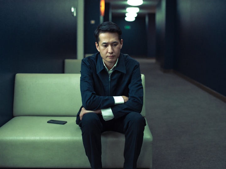 A man in a jacket sitting alone on a bench in a hotel corridor, phone face down beside him