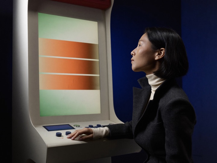 A woman in a charcoal blazer seated at a gaming cabinet, one hand on its controls, lit by the glow of its display