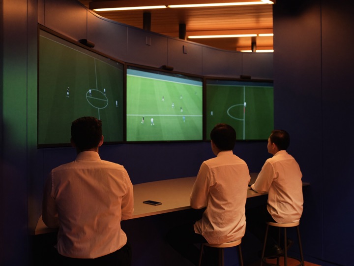A sports lounge wall of large screens showing a soccer pitch, three guests on bar stools